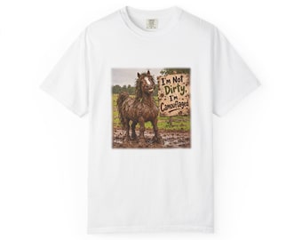 May include: A white short-sleeved t-shirt featuring a graphic print of a brown horse covered in mud standing in a field. Next to the horse is a wooden sign with the text "I'm Not Dirty, I'm Camouflaged" printed in dark brown lettering.