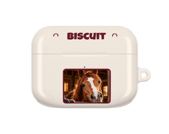 Op de afbeelding: Een crèmekleurige beschermhoes voor draadloze oordopjes met het woord BISCUIT in donkerrode tekst aan de bovenkant, geflankeerd door twee kleine hoefijzerpictogrammen. Een rechthoekige afbeelding van een bruin paard met een witte bles is gecentreerd.