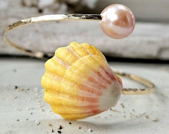 May include: A gold-toned open cuff bracelet featuring a hammered metal band. One end of the bracelet is adorned with a single, smooth, pale pink pearl, while the other end features a textured, yellow and orange seashell with distinct ridges.
