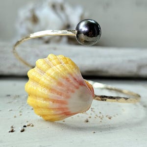 May include: A gold-toned open cuff bracelet featuring a hammered metal band. One end of the bracelet is adorned with a small, dark metallic sphere, while the other end features a natural seashell with yellow and soft pink coloration.