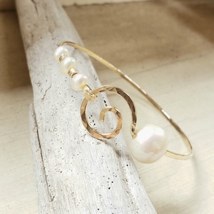 Puede incluir: Brazalete dorado con diseño de espiral y una perla grande y redonda. El brazalete también incluye tres perlas más pequeñas. El metal tiene una textura martillada. Una joya delicada.