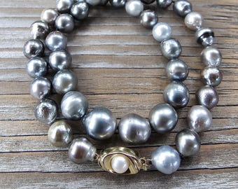 May include: A necklace composed of large, lustrous, gray pearls. The pearls vary in size and shade, with a mix of silver, charcoal, and iridescent tones. The clasp is gold-toned, with a single white pearl at its center. The necklace is displayed on a wooden surface.
