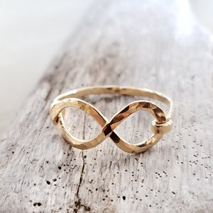May include: A gold infinity ring with a hammered texture. The ring is on a light brown wooden surface.