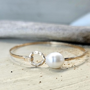 May include: A gold-colored bangle bracelet with a large, round, white pearl. The bracelet features a hammered texture and a unique clasp design. The pearl is the focal point of the bracelet.