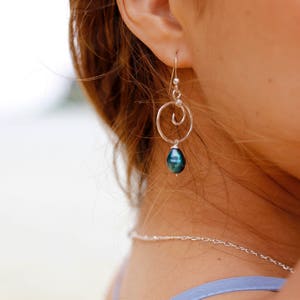 May include: A silver earring with a dark blue-green pearl pendant. The earring features a small hoop and a spiral design. A silver necklace is also visible.