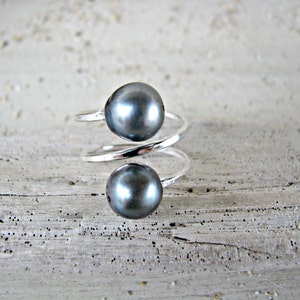 May include: A silver ring with two dark gray pearls. The ring is twisted and has a modern design.