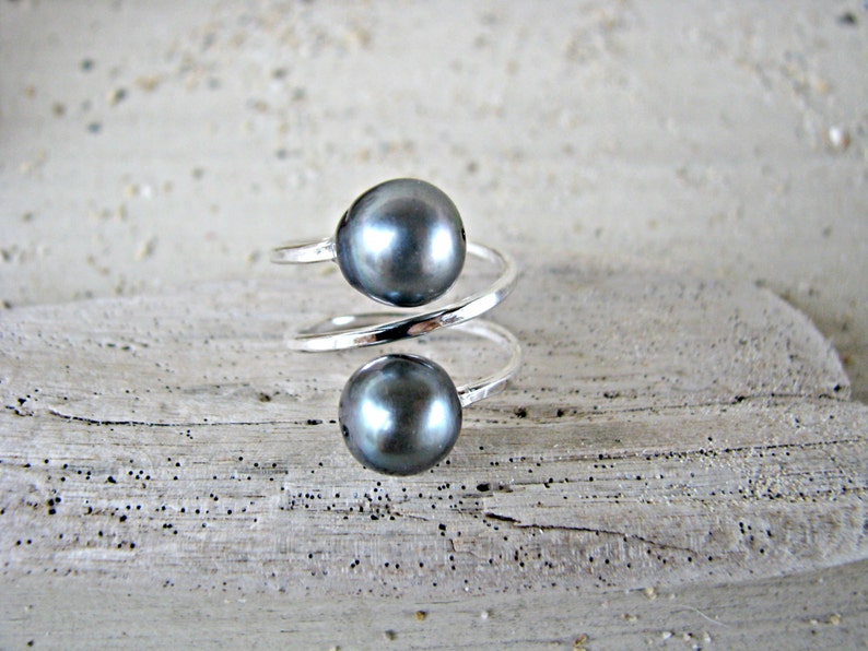 May include: A silver ring with two dark gray pearls. The ring is twisted and has a modern design.