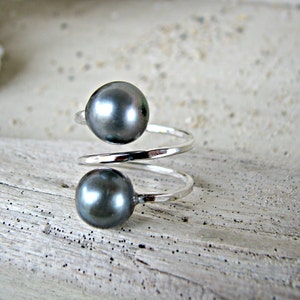 May include: A silver ring with two dark gray pearls. The ring is twisted and has a simple design.