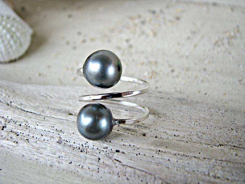 May include: A silver ring with two dark gray pearls. The ring is twisted and has a simple design.