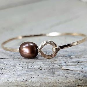 Puede incluir: Un brazalete dorado con textura martillada. El brazalete presenta una gran perla marrón oscuro y un cierre circular. La joya está sobre una superficie de madera envejecida.