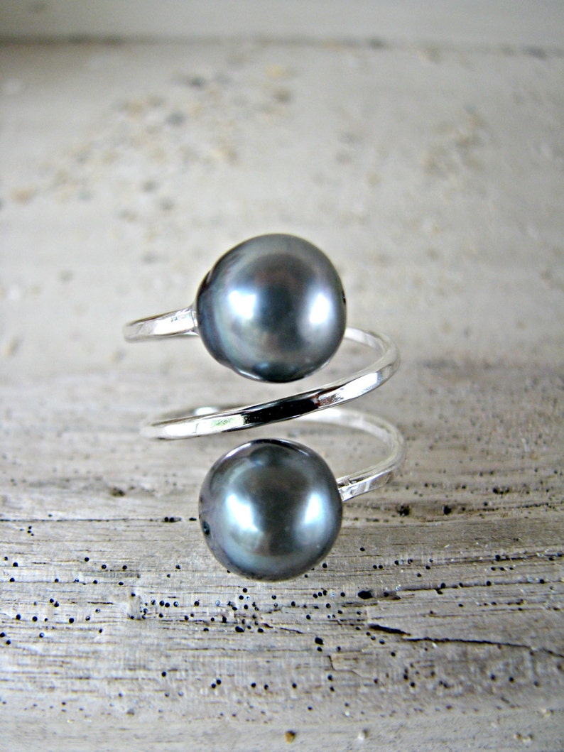 May include: A silver ring with a double spiral design features two dark gray pearls. The ring is on a light gray wooden surface.