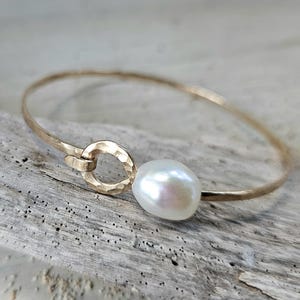 May include: A gold-colored bangle bracelet with a hammered texture and a large, lustrous white pearl. The bracelet features a circular clasp and a simple, elegant design, perfect for everyday wear.