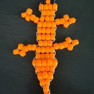 May include: A lizard-shaped figure crafted from bright orange plastic pony beads strung on white cord. The design features two black beads for eyes at the head, a segmented body, and four limbs extending from the sides, ending in a tapered tail.