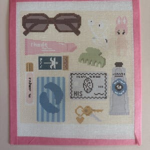 May include: A needlepoint canvas featuring a collection of items including brown sunglasses, a pink tube, a blue striped card, a green hair claw, a credit card, a grey tube, and gold keys, all framed by a pink border on a light background.