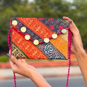 May include: A rectangular envelope-style clutch bag featuring a patchwork of multicolored embroidered fabric panels in shades of orange, red, yellow, and dark blue, accented with gold-toned circular coin charms and a bright pink trim.