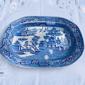 May include: An oval ceramic platter featuring a blue and white willow pattern. The design depicts pagodas, a bridge, trees, and birds. A small white rectangular sticker with the text 374-E is affixed to the upper centre of the rim.