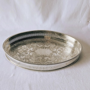 May include: An oval silver-toned serving tray featuring an intricate engraved floral pattern across the base and a raised gallery rim with a vertical slotted design and a beaded edge detail along the outer perimeter of the tray.