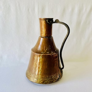 May include: A copper pitcher with a wide, conical base that tapers into a cylindrical neck. The vessel features a dark, curved metal handle attached to the side. The surface displays a warm, metallic finish with visible signs of wear and patina.