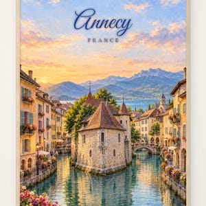 Op de afbeelding: Een ingelijste kunstdruk van het Palais de l'Île in Annecy, Frankrijk, met een stenen gebouw omringd door een kanaal, kleurrijke bloemen op balkons en bergen onder een zonsonderganghemel met de tekst Annecy FRANCE.