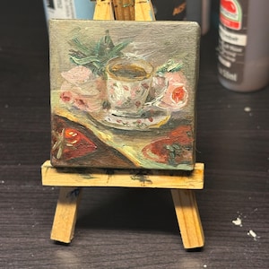 May include: A small square oil painting on canvas displayed on a miniature wooden easel. The painting depicts a floral teacup and saucer surrounded by pink flowers and green leaves, with dark red fruit shapes in the foreground on a muted background.