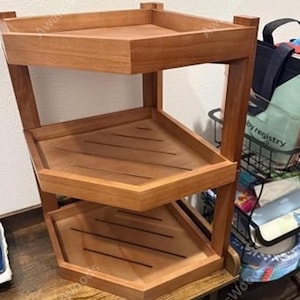 May include: A three-tiered corner shelf constructed from medium-toned wood. The unit features a triangular shape with raised edges on each level. The two lower shelves include horizontal slats, while the top shelf has a solid, flat surface.