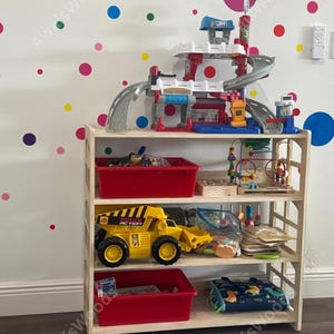 May include: A light-colored wooden four-tier shelving unit filled with various toys, including a multi-level plastic parking garage, a large yellow Maxx Action dump truck, two red plastic storage bins, a bead maze, and assorted wooden puzzles.