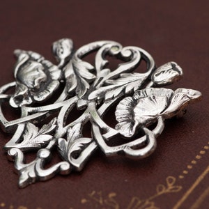 May include: A detailed silver-toned floral brooch featuring stylized flowers and leaves. The brooch has a classic, elegant design with intricate detailing. The metal has a slightly aged appearance, enhancing the vintage aesthetic. The brooch is likely made of a metal alloy.