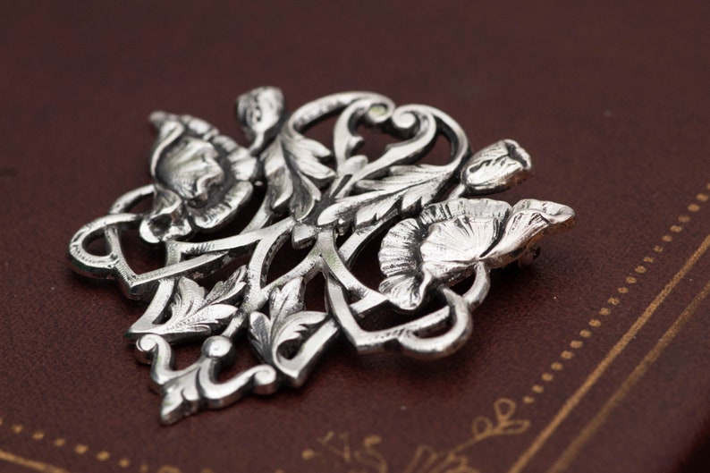 May include: A detailed silver-toned floral brooch featuring stylized flowers and leaves. The brooch has a classic, elegant design with intricate detailing. The metal has a slightly aged appearance, enhancing the vintage aesthetic. The brooch is likely made of a metal alloy.