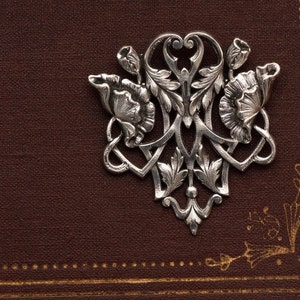 May include: A silver-colored floral brooch with intricate details, including stylized flowers and leaves. The brooch is set against a textured, dark brown background with gold accents. The design is symmetrical and ornate.