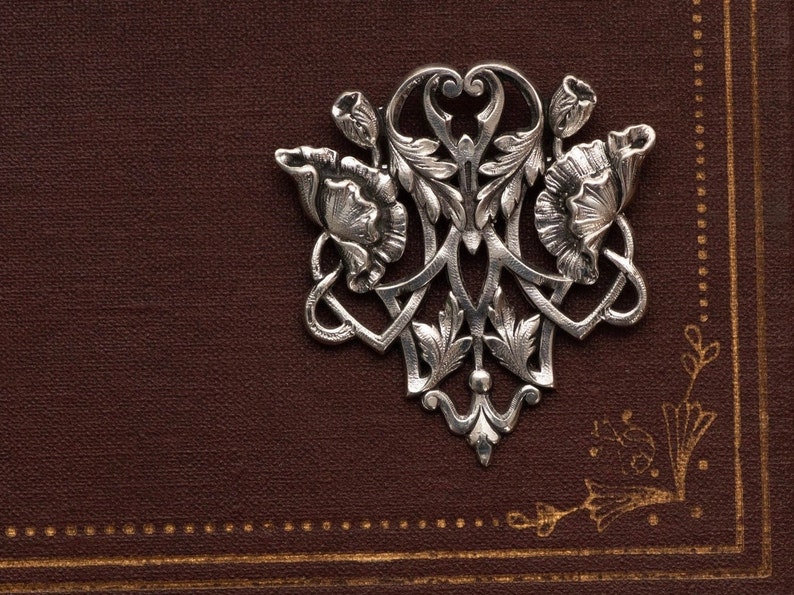 May include: A silver-colored floral brooch with intricate details, including stylized flowers and leaves. The brooch is set against a textured, dark brown background with gold accents. The design is symmetrical and ornate.