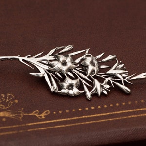 May include: A silver-toned floral brooch featuring a detailed design of flowers and leaves. The brooch is a botanical-themed accessory, perfect for adding a touch of nature-inspired elegance to any outfit. The brooch is displayed on a brown surface.