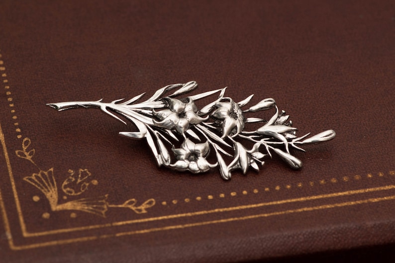 May include: A silver-toned floral brooch featuring a detailed design of flowers and leaves. The brooch is a botanical-themed accessory, perfect for adding a touch of nature-inspired elegance to any outfit. The brooch is displayed on a brown surface.