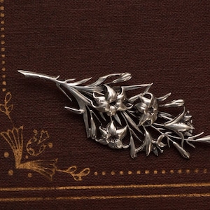 May include: A silver-toned floral brooch featuring detailed lily blossoms and leaves. The brooch is set against a dark brown background, creating a contrast that highlights the intricate design of the jewelry.