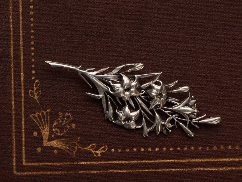 May include: A silver-toned floral brooch featuring detailed lily blossoms and leaves. The brooch is set against a dark brown background, creating a contrast that highlights the intricate design of the jewelry.