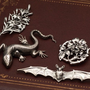 May include: A collection of silver-toned brooches and pendants. The designs include a floral spray, a lizard, a floral wreath, and a bat with outstretched wings. The pieces are displayed on a brown leather surface.