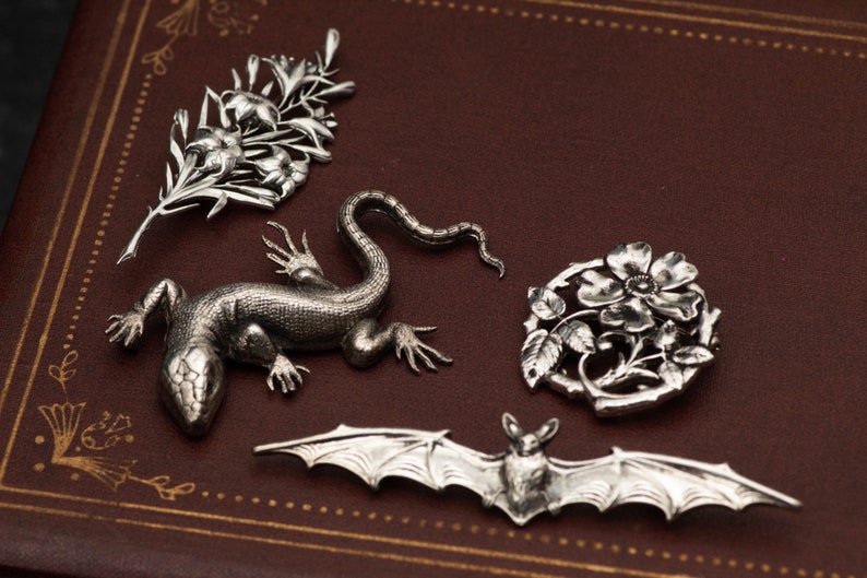 May include: A collection of silver-toned brooches and pendants. The designs include a floral spray, a lizard, a floral wreath, and a bat with outstretched wings. The pieces are displayed on a brown leather surface.
