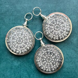 May include: Three silver-colored, round decorative items with intricate floral filigree patterns. Each has a small ring at the top and a decorative stem. The items are arranged on a teal-colored surface.