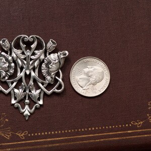 May include: A silver-toned floral brooch with intricate details, featuring stylized flowers and leaves. A U.S. quarter coin is placed next to the brooch for size comparison. The brooch is set against a dark brown background.