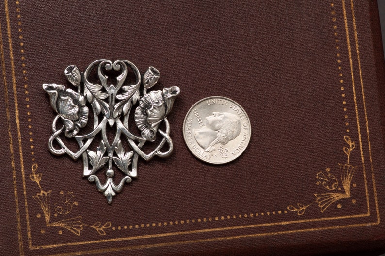 May include: A silver-toned floral brooch with intricate details, featuring stylized flowers and leaves. A U.S. quarter coin is placed next to the brooch for size comparison. The brooch is set against a dark brown background.