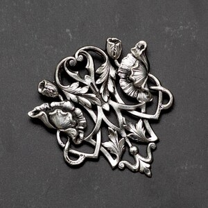 May include: A silver-toned floral brooch featuring a detailed design of stylized flowers, buds, and leaves. The brooch has an Art Nouveau style with flowing lines and intricate details. The brooch is set against a dark gray background.