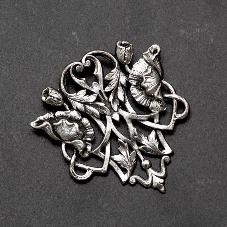 May include: A silver-toned floral brooch featuring a detailed design of stylized flowers, buds, and leaves. The brooch has an Art Nouveau style with flowing lines and intricate details. The brooch is set against a dark gray background.