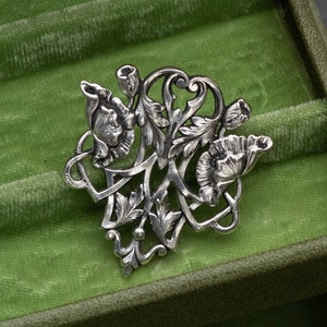 May include: A silver-toned floral brooch featuring stylized flowers, buds, and leaves. The brooch has an intricate, Art Nouveau design with flowing lines and detailed textures. The brooch is displayed on a green velvet surface.