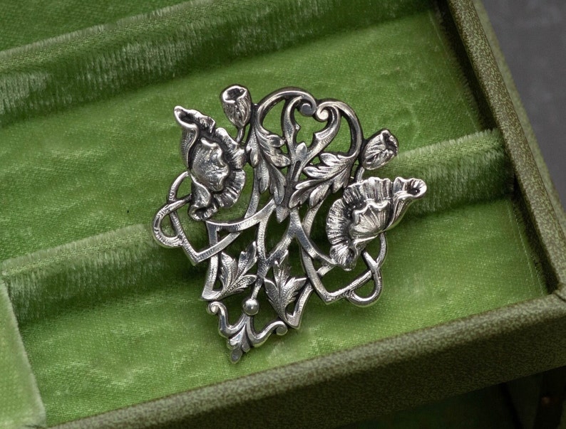 May include: A silver-toned floral brooch featuring stylized flowers, buds, and leaves. The brooch has an intricate, Art Nouveau design with flowing lines and detailed textures. The brooch is displayed on a green velvet surface.