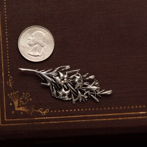 May include: A silver-colored floral brooch with detailed flower and leaf design. A U.S. quarter coin is next to the brooch for size comparison. The brooch is on a brown, textured surface.