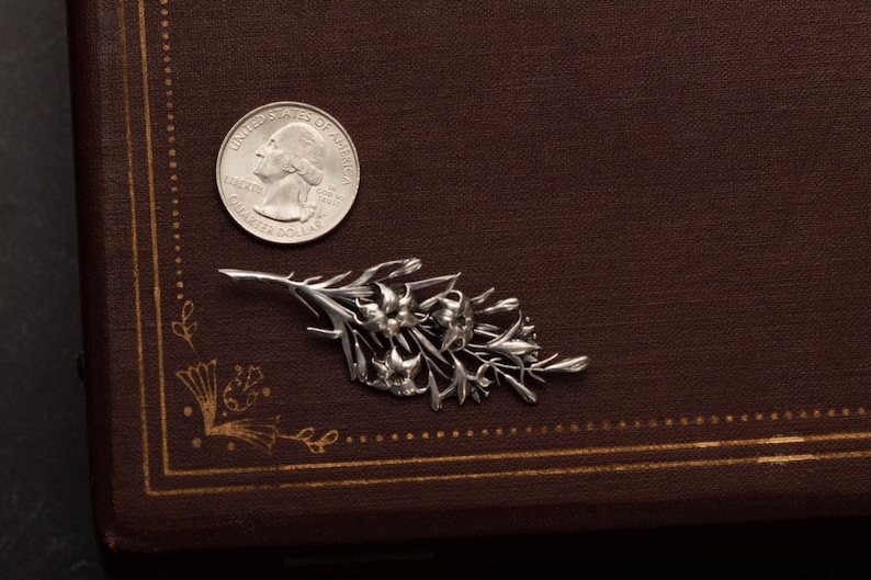 May include: A silver-colored floral brooch with detailed flower and leaf design. A U.S. quarter coin is next to the brooch for size comparison. The brooch is on a brown, textured surface.