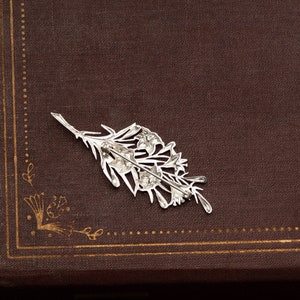 May include: A silver-colored floral brooch, shaped like a sprig of flowers, rests on a brown surface. The brooch has intricate details and a pin backing. The background is a dark brown, possibly leather-bound book.