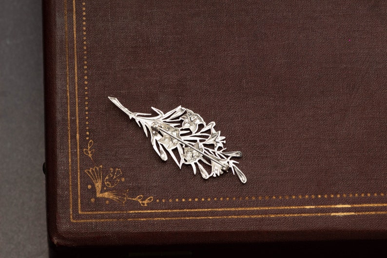 May include: A silver-colored floral brooch, shaped like a sprig of flowers, rests on a brown surface. The brooch has intricate details and a pin backing. The background is a dark brown, possibly leather-bound book.