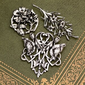 May include: Three silver-toned floral brooches with intricate details are displayed on a green surface. The brooches feature various flower and leaf designs, including a circular arrangement, a sprig, and a stylized floral shape. The background has a decorative border.