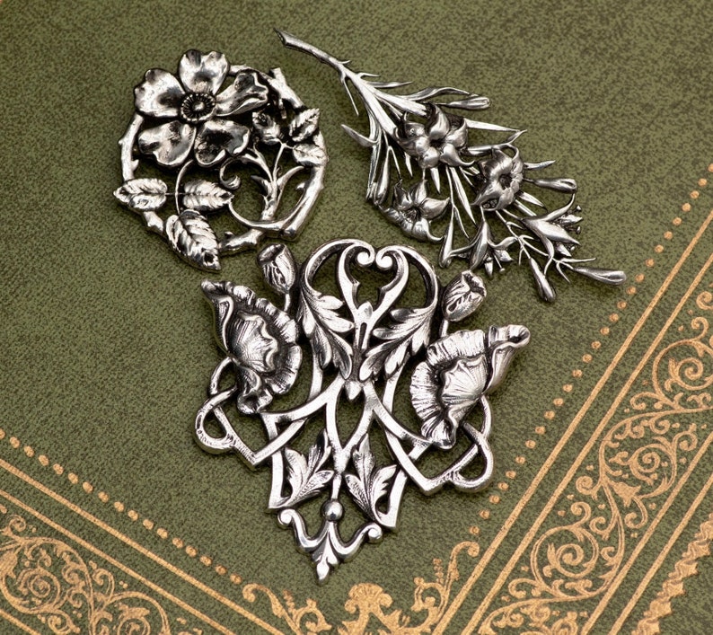 May include: Three silver-toned floral brooches with intricate details are displayed on a green surface. The brooches feature various flower and leaf designs, including a circular arrangement, a sprig, and a stylized floral shape. The background has a decorative border.