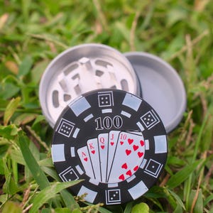 May include: A circular metal herb grinder featuring a black and white poker chip design on the lid. The lid displays a royal flush of hearts, the number 100, and dice patterns. The interior shows silver metal teeth for grinding herbs.